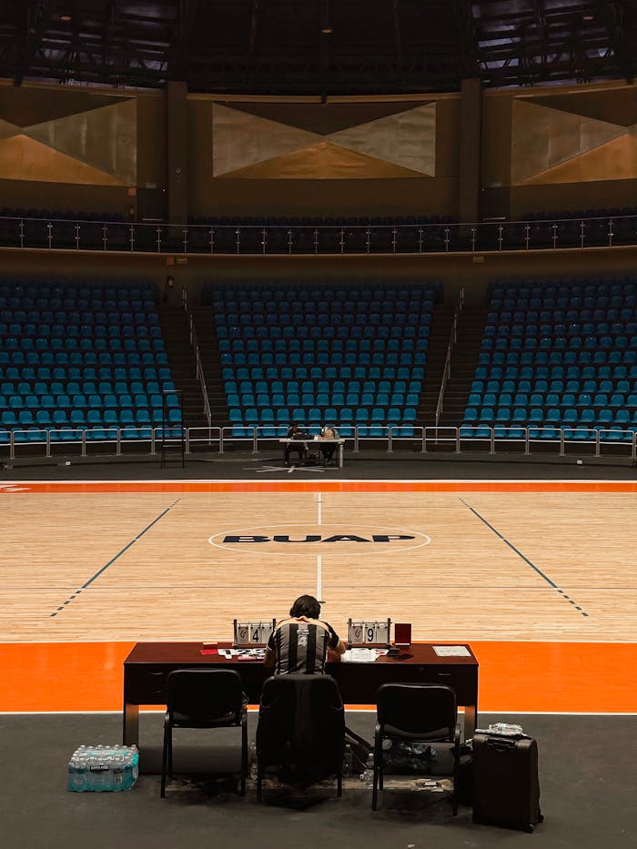 A lone official at a table on an empty basketball court with empty blue seats.